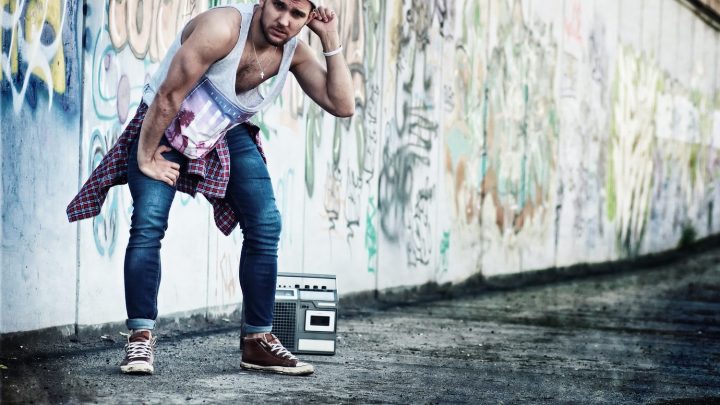 Des artistes hip-hop se produisent devant un mur couvert de graffitis, avec un DJ aux platines et un danseur au premier plan.