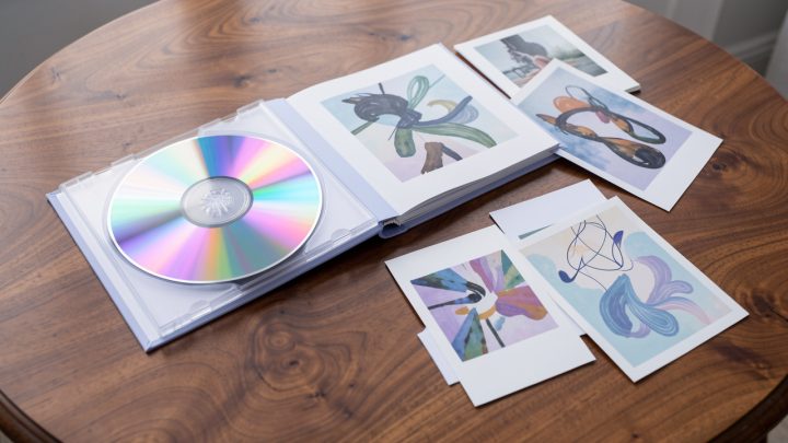 Album musical collector ouvert sur une table, avec un CD coloré, un photobook, des photocards décoratives et un poster artistique, éclairé par une lumière douce.
