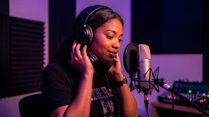 Une jeune chanteuse R&B dans un home studio intimiste, casque autour du cou, près d’un microphone de studio, baignée dans une lumière chaude violette et bleue.