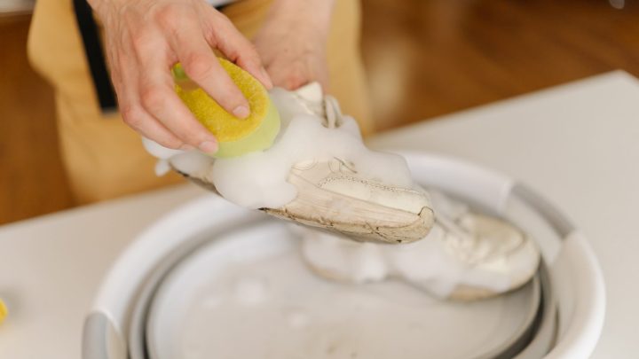 Personne nettoyant une paire de sneakers blanches avec une brosse et de l’eau savonneuse à la maison.